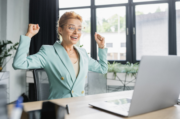 allegra donna d'affari rossa in occhiali che mostra il gesto di vittoria vicino al computer portatile in ufficio - Foto, immagini