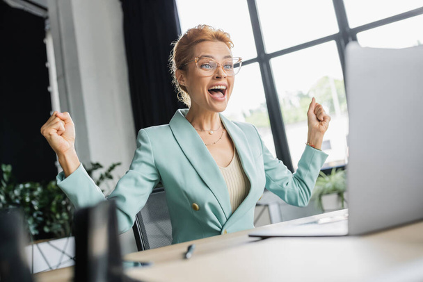 overjoyed businesswoman in eyeglasses shouting and showing win gesture near blurred laptop in office - Photo, Image