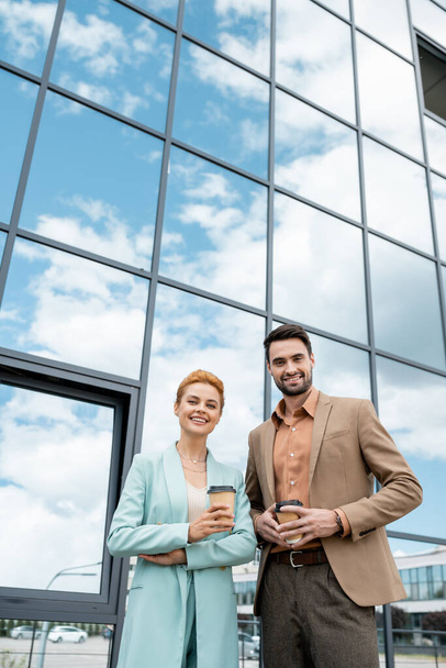 stylish business people with paper cups smiling at camera near modern building with glass facade - Foto, Imagen