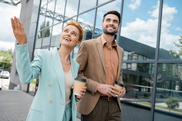 glückliche rothaarige Geschäftsfrau, die wegschaut und mit der Hand zeigt, während sie mit einem Kollegen auf der Stadtstraße spaziert - Foto, Bild