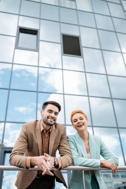 fröhliche und trendige Manager lächeln in die Kamera nahe Geländer und Stadtgebäude mit Glasfassade - Foto, Bild