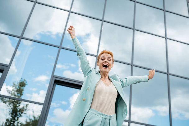 low angle view of excited businesswoman showing triumph gesture and screaming near glass building in city - Fotografie, Obrázek