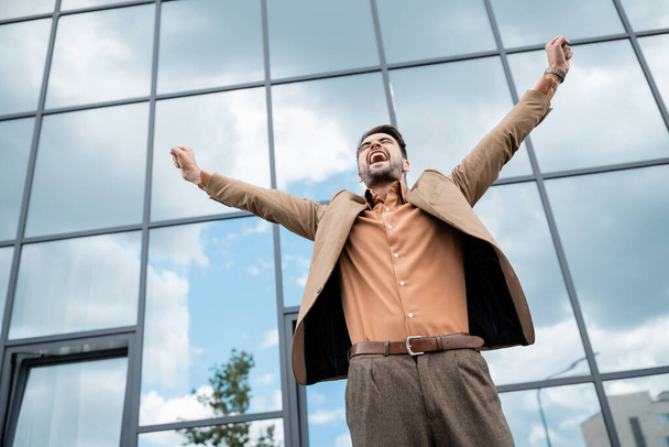 низкий угол обзора успешного сверхрадостного бизнесмена в модной одежде, показывающей жест триумфа возле современного городского здания  - Фото, изображение