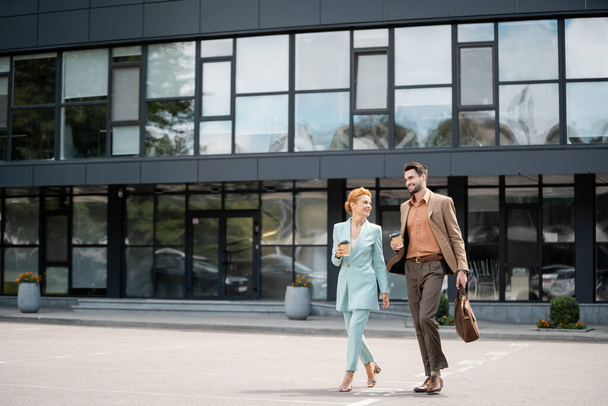 full length of trendy business partners with paper cups walking near modern building on urban street - Zdjęcie, obraz