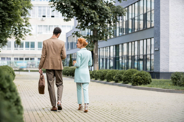 vista trasera de la mujer de negocios feliz y elegante hablando con su colega mientras camina a lo largo de edificios modernos en la ciudad - Foto, Imagen