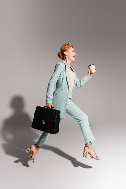 full length of amazed businesswoman in stylish suit levitating with coffee to go and black briefcase on grey background - Fotografie, Obrázek