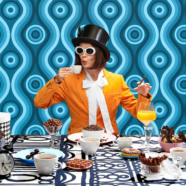 Artistic woman in stylish top hat and sunglasses sitting at table with many chocolate sweets and drinking coffee. Pattern blue background. Concept of pop art photography, creativity, food, inspiration - Zdjęcie, obraz