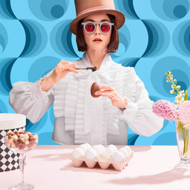 Chocolates lover. Stylish woman in cylinder har and sunglasss eating chocolate eater egg. Blue pattern background. Concept of pop art, creativity, food, inspiration and imagination, contemporary photo - Fotoğraf, Görsel