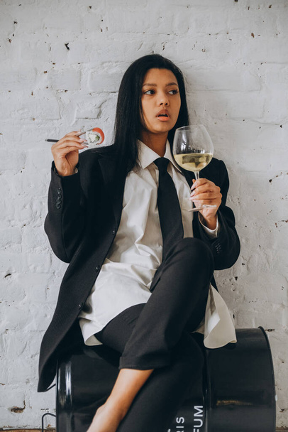 A dark-skinned young woman sits pretentiously in a white room and eats sushi with chopsticks. African American woman in a business suit eats rolls. - Photo, Image