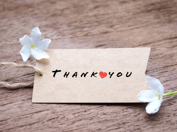 Słowo "dziękuję" i małe czerwone serce na brązowym tagu i biały kwiat na rustykalnym drewnianym tle. - Zdjęcie, obraz