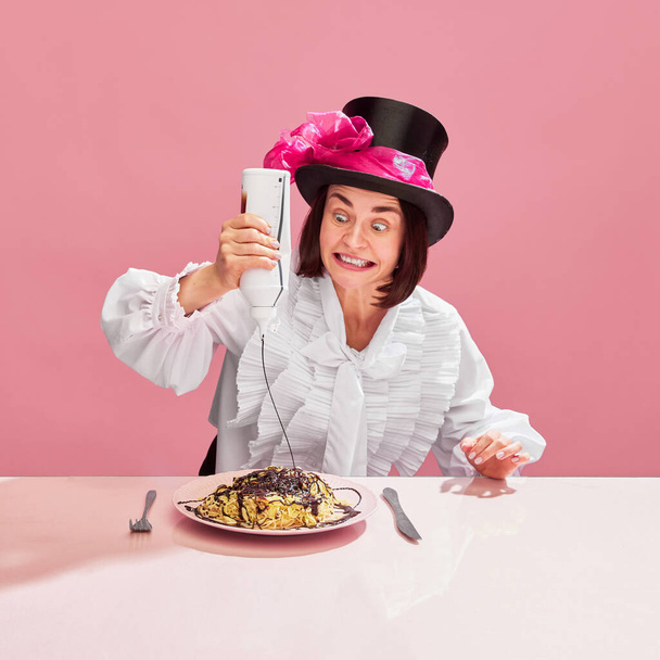 Crazy face. Emotive woman in cylinder hat emotionally squeezing chocolate sauce on spaghetti over pink background. Sweets lover. Concept of pop art, creativity, food, inspiration, emotions, meme look - Fotó, kép
