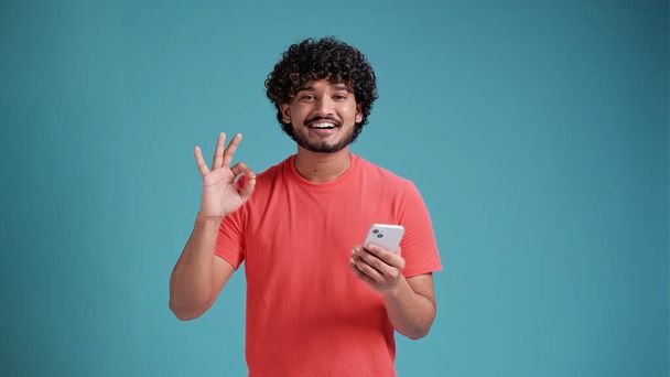 Young indian man using smartphone doing ok sign with fingers, excellent symbol in coral t-shirt on blue studio background. - Fotografie, Obrázek