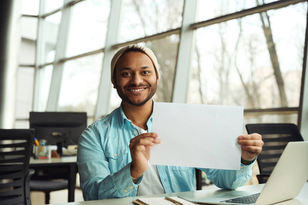 Szczęśliwy człowiek w kapeluszu siedzi przy stole z kawałkiem papieru w rękach w centrum biznesowym - Zdjęcie, obraz