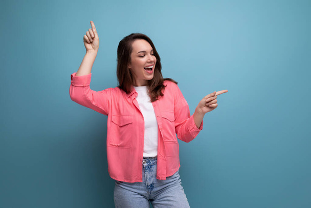 vrolijk brunette dame in een roze shirt en jeans wijst haar vinger naar de infomatie op een studio achtergrond. - Foto, afbeelding