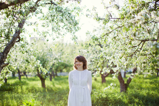 Beautiful caucasian woman in white dress walks in spring blossom garden - Фото, изображение