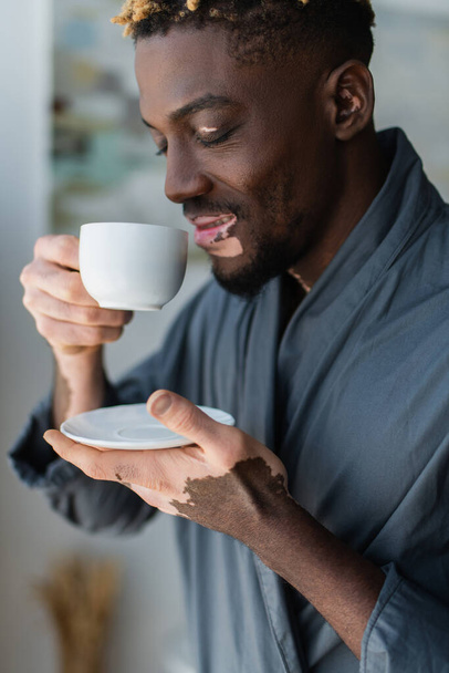 Joyful african american man with vitiligo holding cup of coffee at home  - Fotografie, Obrázek