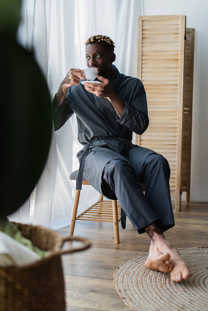 African american man with vitiligo in robe drinking coffee on bedroom bench at home  - Фото, изображение