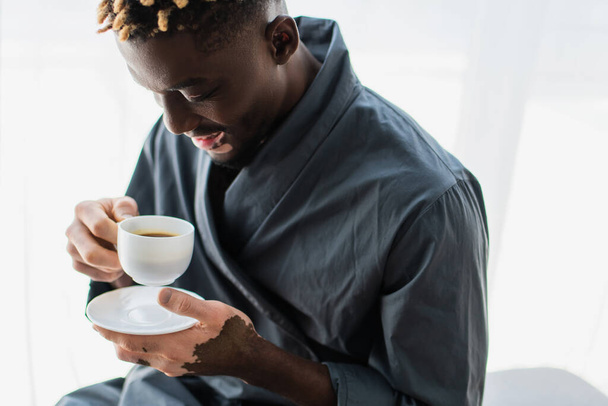 Carefree african american man with vitiligo holding cup of coffee at home  - Zdjęcie, obraz