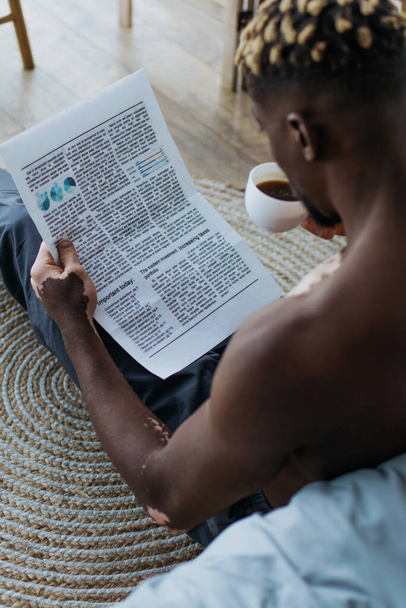 Shirtless african american man with vitiligo reading newspaper and drinking coffee at home  - Foto, immagini