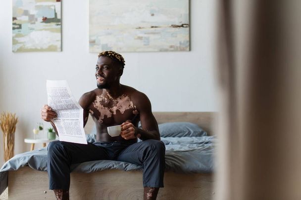 Carefree african american man with vitiligo holding coffee cup and newspaper while sitting on bed at home  - Photo, image