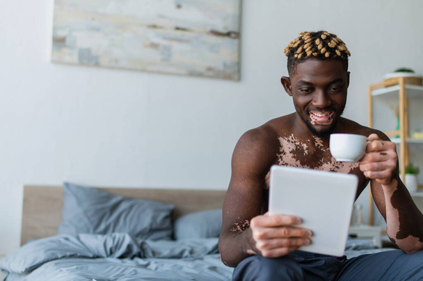 Shirtless african american man with vitiligo smiling while using digital tablet and holding cup of coffee in bedroom  - Фото, зображення