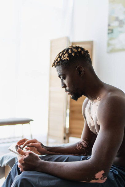 Вид збоку без сорочки афроамериканський чоловік з вітіліго, використовуючи цифровий планшет, сидячи на ліжку вдома
  - Фото, зображення