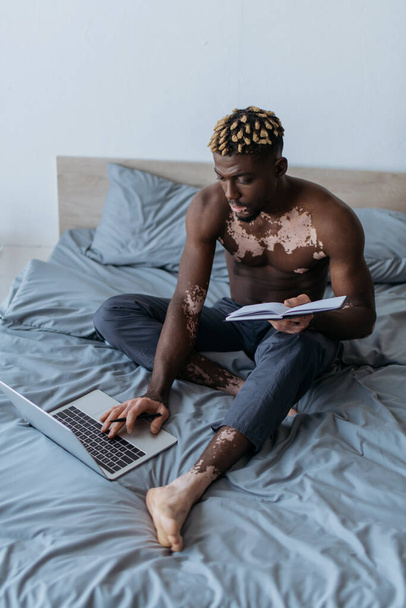 Shirtless african american freelancer with vitiligo holding notebook and using laptop in bedroom  - Foto, Imagem