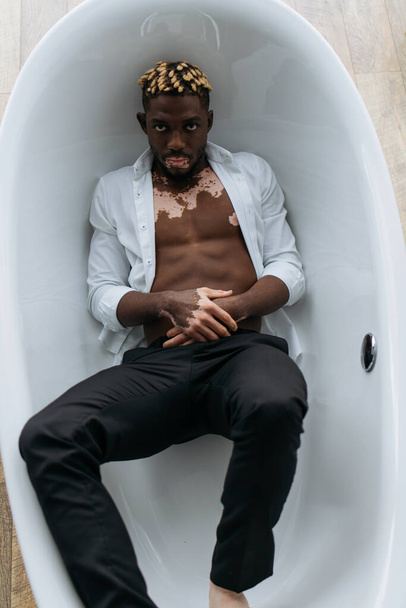 Top view of african american man with vitiligo in shirt and pants lying in bathtub at home  - Fotografie, Obrázek