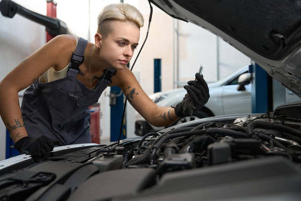 Hardworking female employee enthusiastically looking at detail while repairing car in service center - Photo, Image
