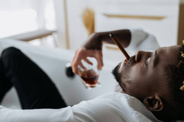 Vista lateral del hombre afroamericano con vitiligo fumando cigarro y sosteniendo whisky borroso en la bañera  - Foto, imagen