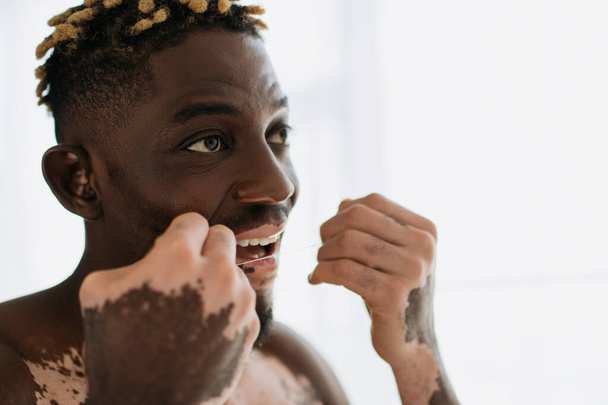 Shirtless african american man with vitiligo using dental floss at home  - Фото, зображення