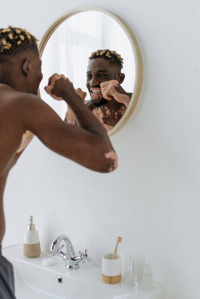 Shirtless african american man with vitiligo flossing teeth near mirror in bathroom  - 写真・画像
