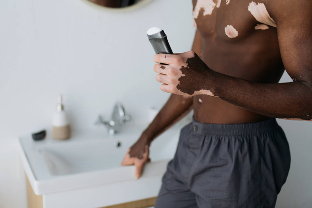 Cropped view of muscular african american man with vitiligo holding antiperspirant in bathroom  - Φωτογραφία, εικόνα