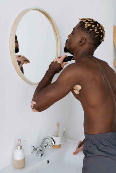Vista laterale di uomo africano americano con la rasatura vitiligine con rasoio elettrico in bagno  - Foto, immagini
