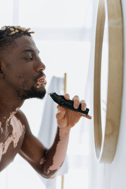 Африканский мужчина без рубашки с витилиго с электрической бритвой возле зеркала в ванной комнате  - Фото, изображение