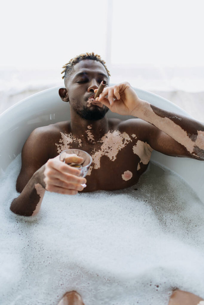 Αφροαμερικάνος με λεύκη καπνίζει πούρο και κρατάει ουίσκι ενώ κάνει μπάνιο στο σπίτι  - Φωτογραφία, εικόνα