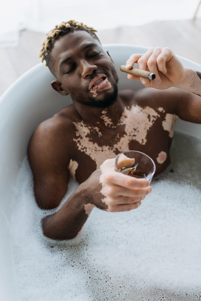 High angle view of african american man with vitiligo holding cigar and whisky while taking bath with foam at home  - Foto, Imagem