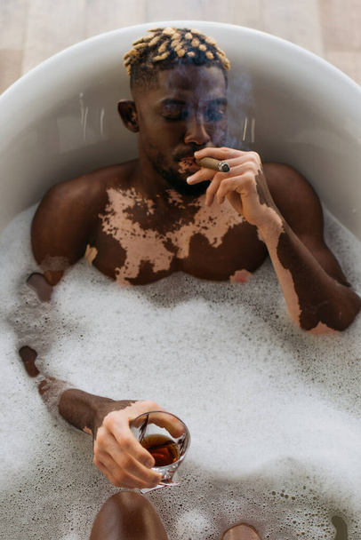 Vista superior do homem afro-americano com vitiligo fumar charuto e segurando uísque no banho com espuma  - Foto, Imagem