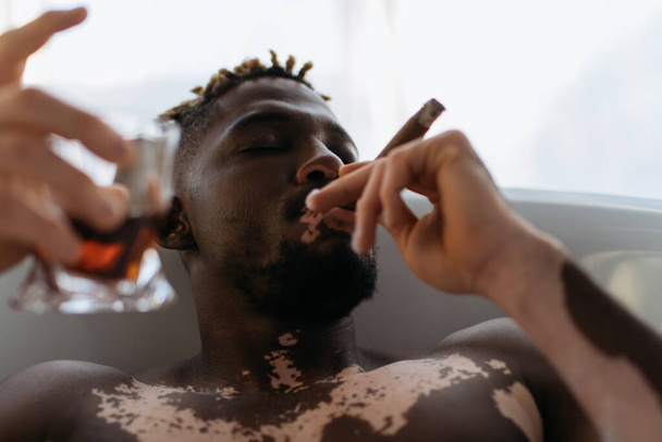 Joven afroamericano con vitiligo fumando cigarro y sosteniendo whisky borroso mientras toma un baño en casa  - Foto, Imagen