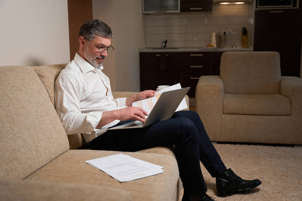Man in business clothes sitting on the sofa, holding laptop and reading documents and working - Foto, imagen