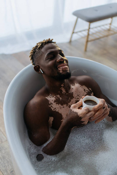High angle view of smiling african american man with vitiligo using earphone and holding coffee in bathtub  - 写真・画像