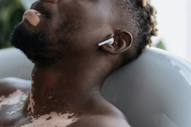 Vista cortada do homem americano africano com vitiligo usando fone de ouvido enquanto toma banho em casa  - Foto, Imagem