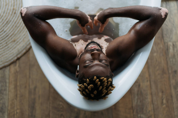 Overhead view of african american man with vitiligo listening music and taking bath at home  - Φωτογραφία, εικόνα