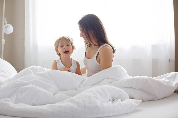 Beautiful blond child and his mother, playing at home in the morning in bed, smiling, laughing and having fun together - Foto, afbeelding