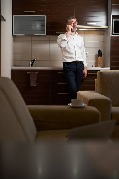 İş kıyafetleri içinde yetişkin bir erkek otel odasında müşteriyle telefonda konuşuyor, çalışıyor. - Fotoğraf, Görsel