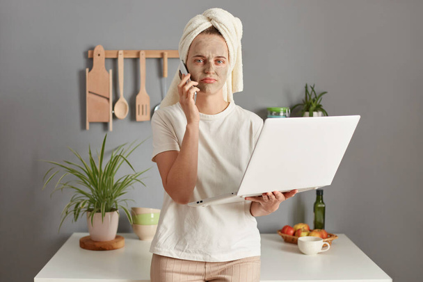 Disappointed displeased young woman posing in kitchen with towel on head working online having troubles on work holding notebook talking phone with sad expression. - Photo, Image