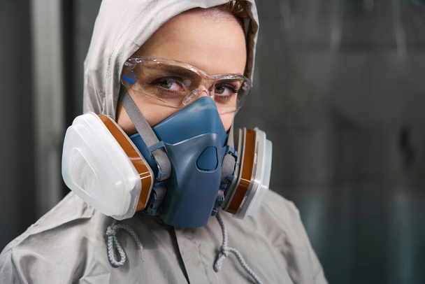Close up view of woman in respirator and goggles looking at camera with serious facial expression indoors - 写真・画像