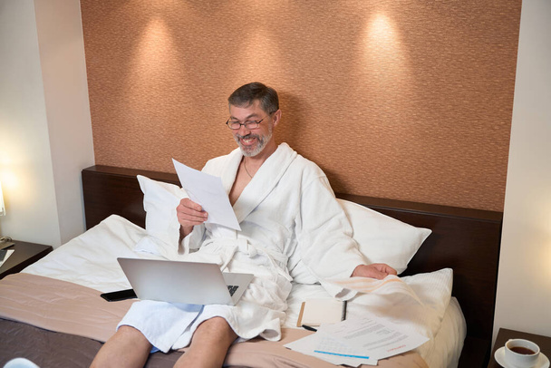 Varón en albornoz sentado en la cama, sosteniendo la computadora portátil y leyendo documentos en el hotel - Foto, imagen