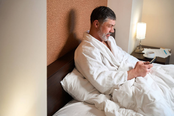 Mężczyzna w szlafroku siedzi na łóżku, trzyma telefon komórkowy, ogląda wiadomości - Zdjęcie, obraz