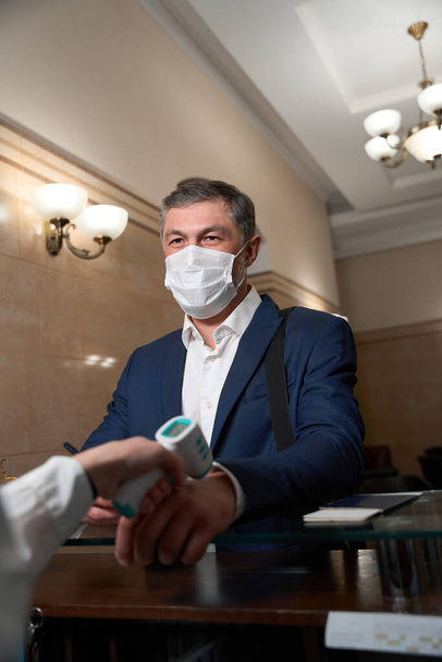 Adult male in business clothes and protective mask standing near reception area, administrator measuring client temperature - Fotoğraf, Görsel